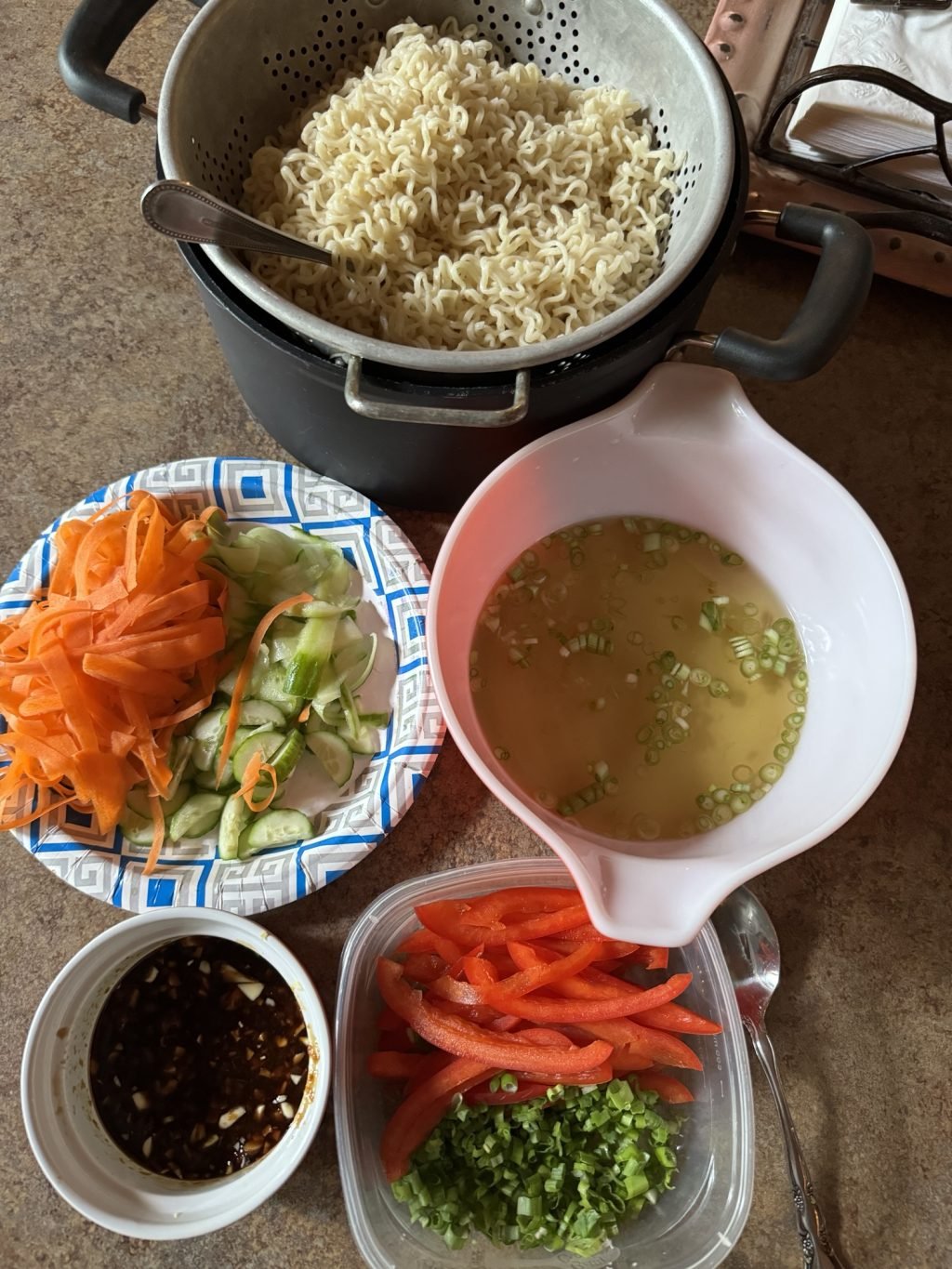 Ramen noodles in a strainer with a fork on on a kitchen counter along with the other ingredients for the cold ramen noodle salad including dashi base, veggies, and sauce.