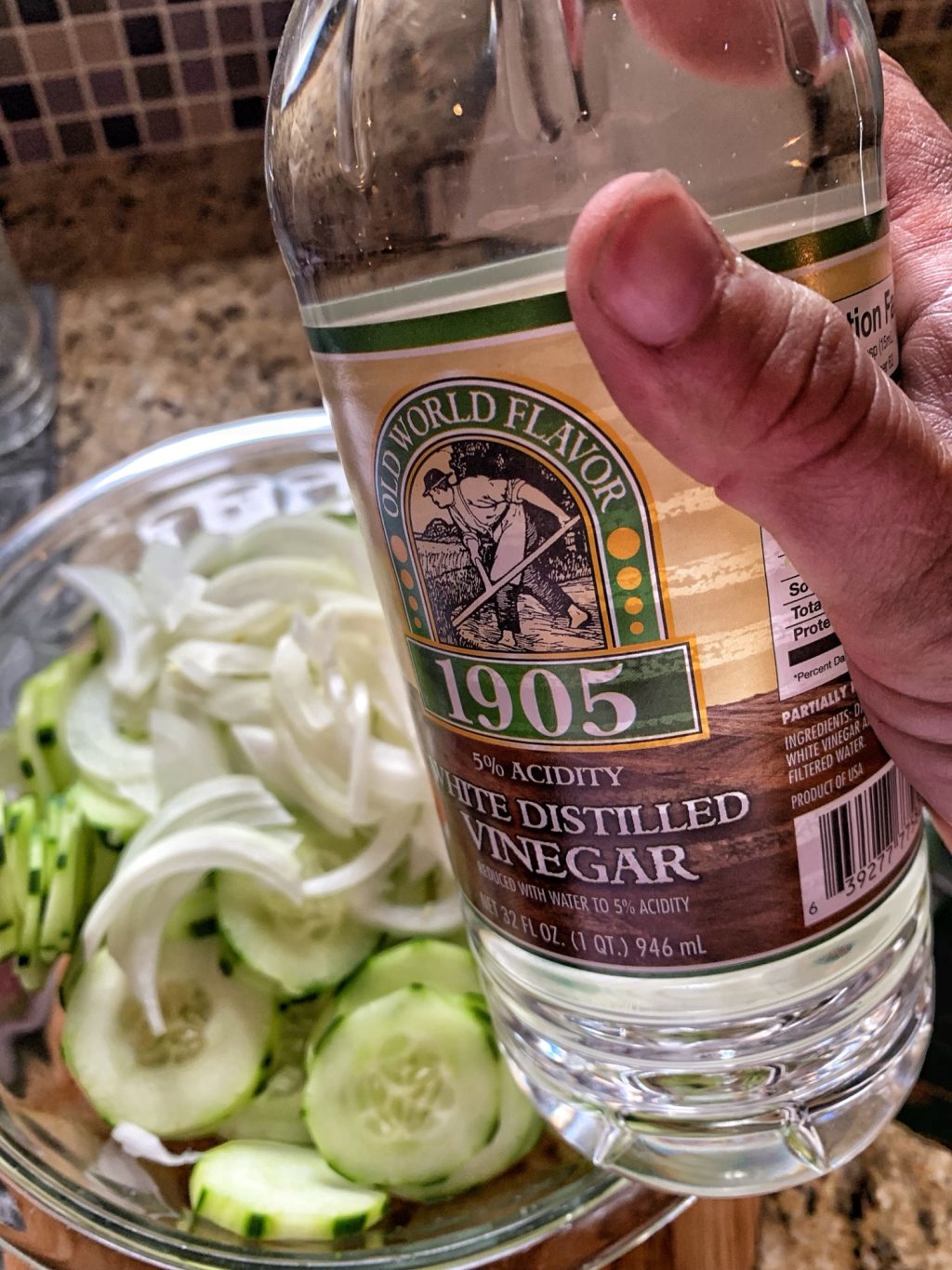 A bottle of white vinegar held over a glass bowl of cucumbers and onion