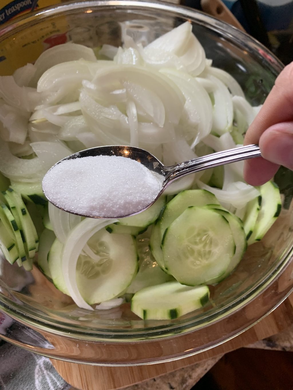 A spoon of sugar held over a glass bowl with sliced cucumbers and onion