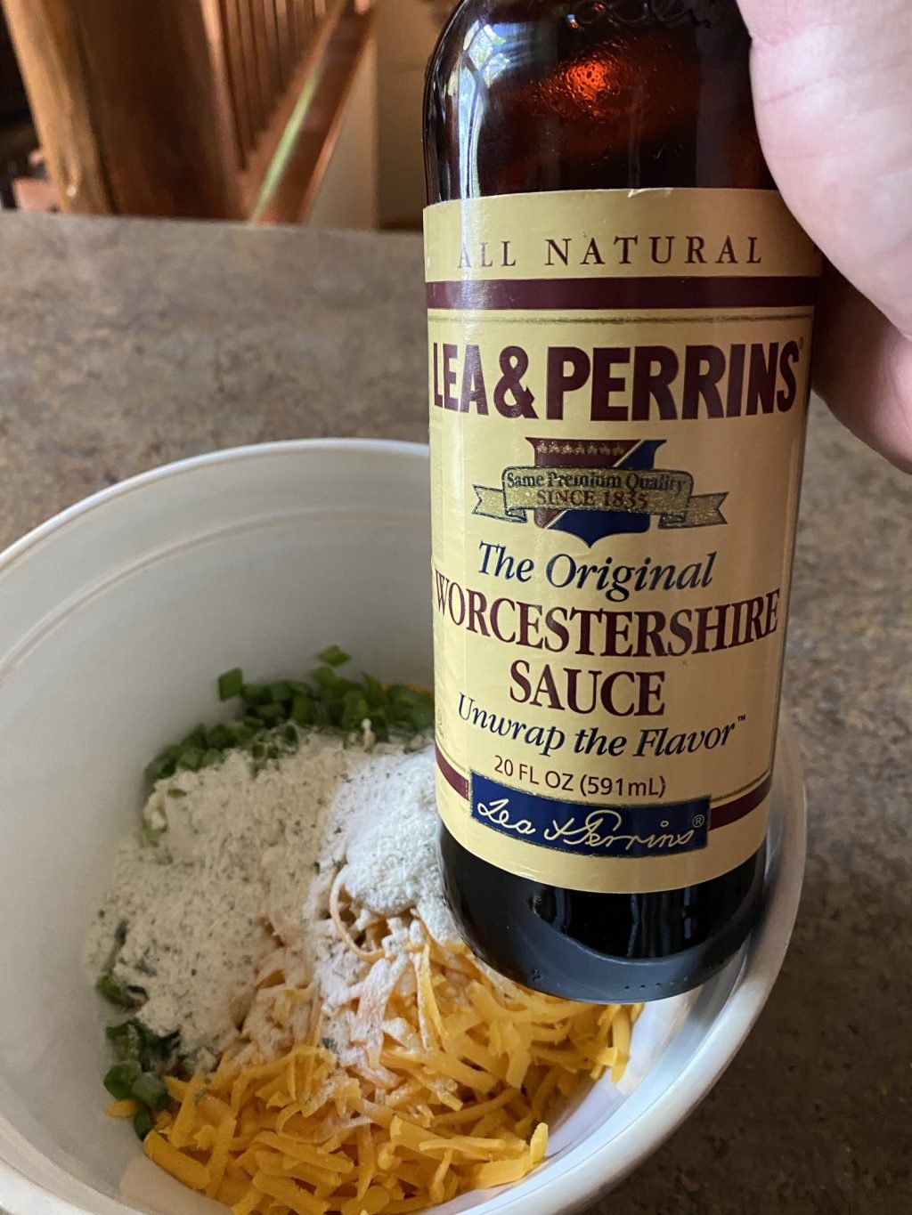 A bottle of Worcestershire sauce held over a bowl with shredded cheese, green onions, and seasoning