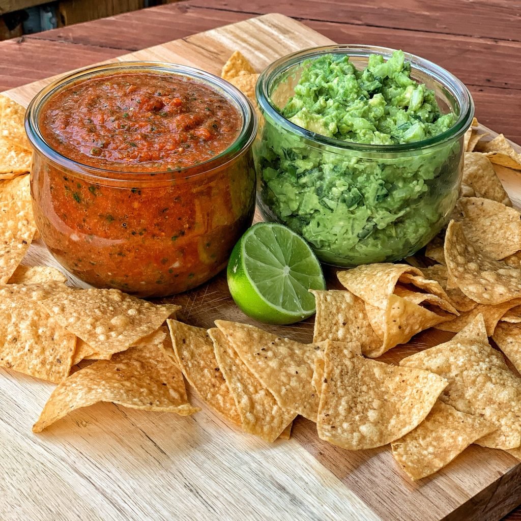 A jar of salsa next to a jar of guacamole surrounded by corn chips and half a lime