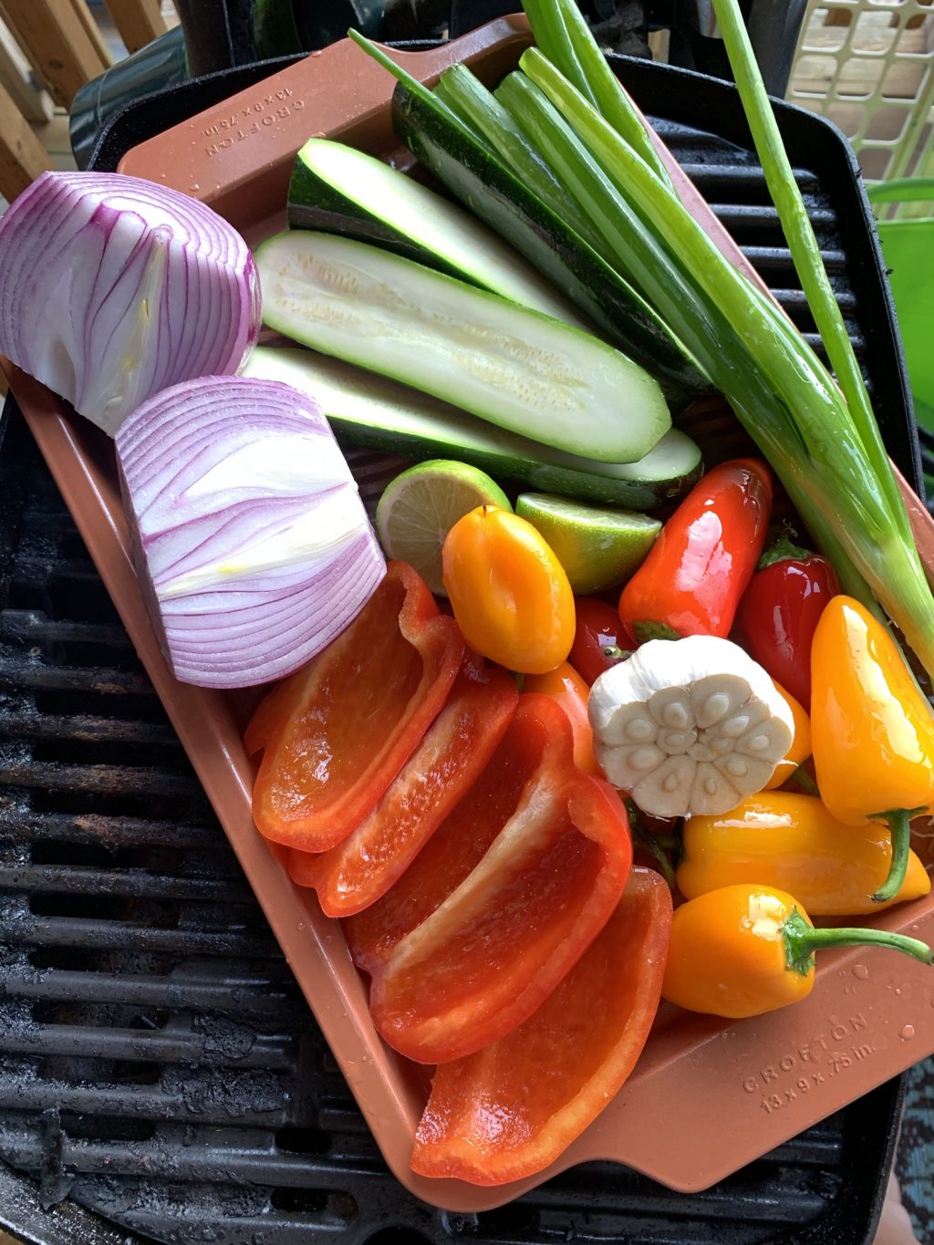 Sliced onion, zucchini, garlic, green onion, and red and orange peppers in a brown container on the grill