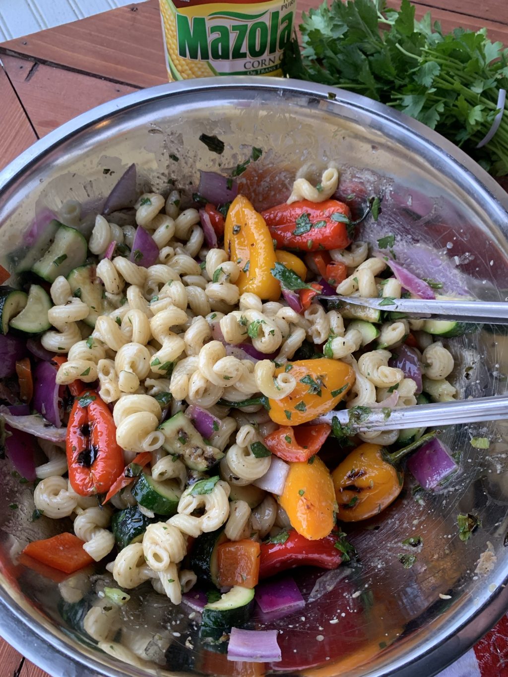 The pasta and grilled vegetables mixed in a metal bowl with tongs for tossing