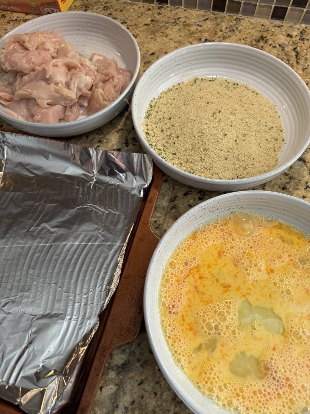 A kitchen counter with three white bowls with raw chicken cutlets, breading mixture, an egg bath, and a baking sheet