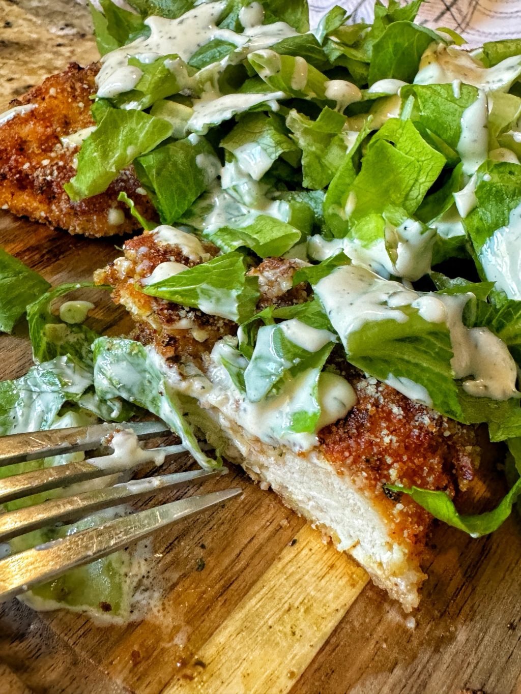 A sliced, breaded chicken cutlet next to a fork topped with lettuce and Caesar salad