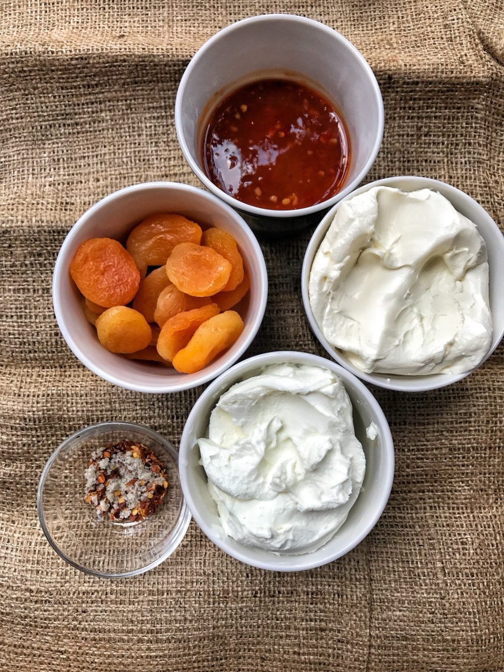 Five containers on burlap with chili sauce, cream cheese, whipped goat cheese, dried apricots, and spices