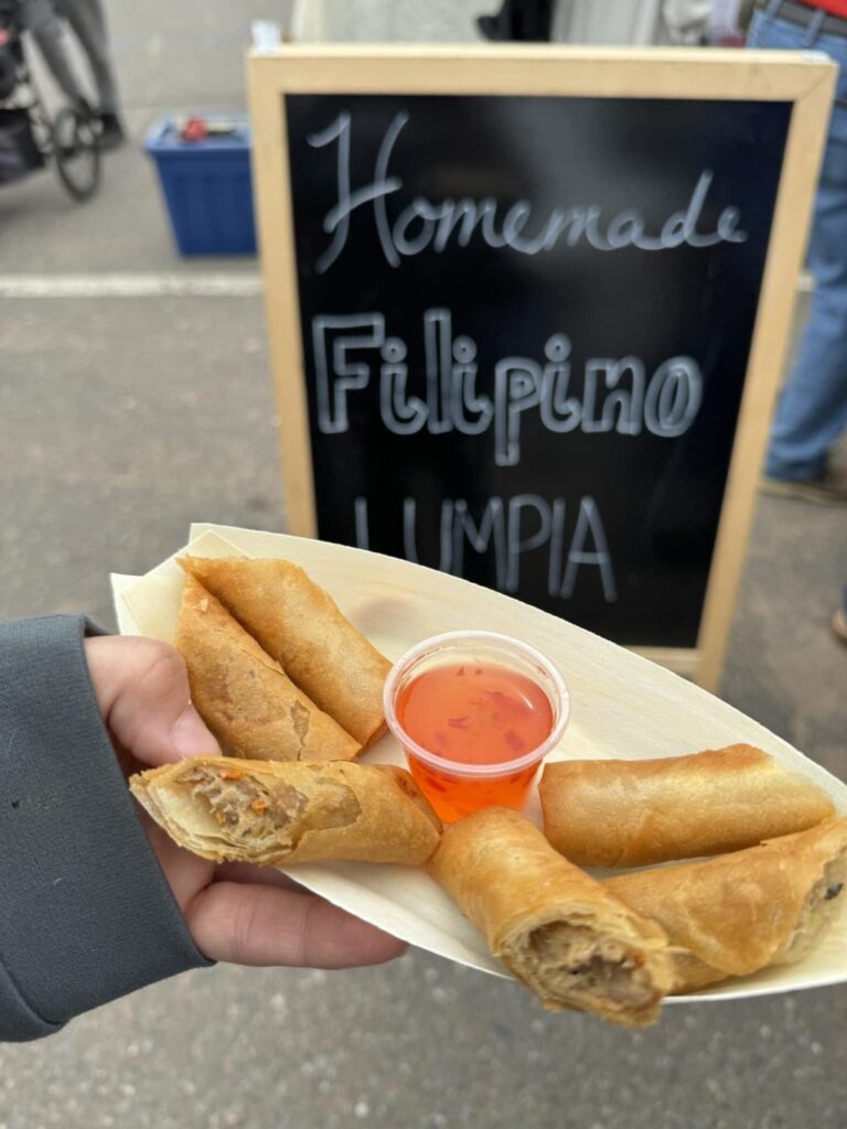 A tray of lumpia spring rolls with a cup of orange sauce I'm holding in a white tray in front of a sign with "Homemade Filipino Lumpia" written on it in white chalk.