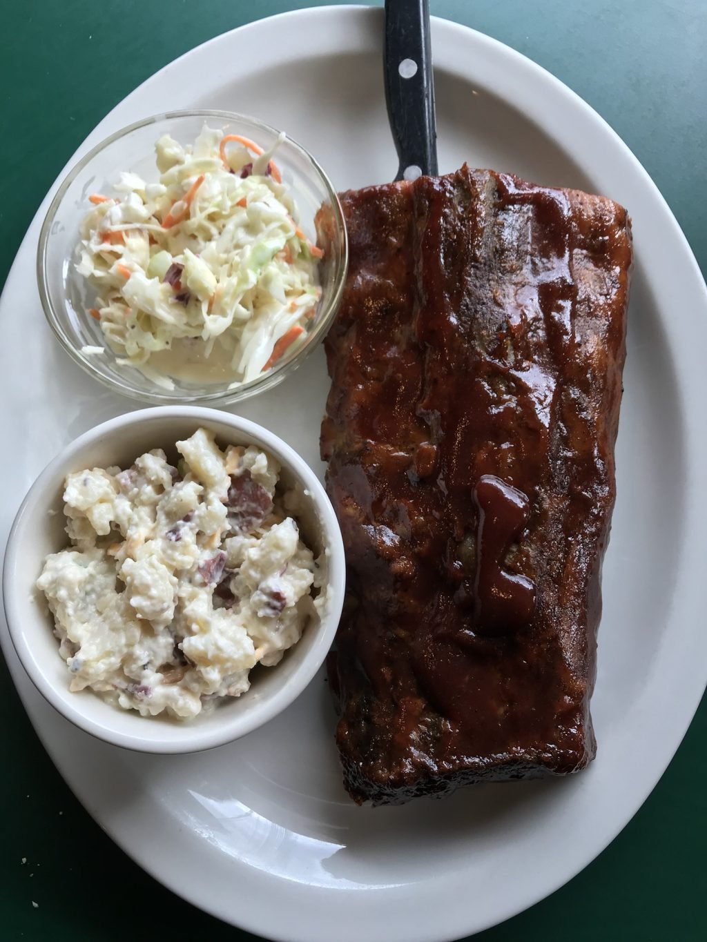 A rack of saucy ribs on a white plate with a knife and a side of cole slaw and another side of potato salad.