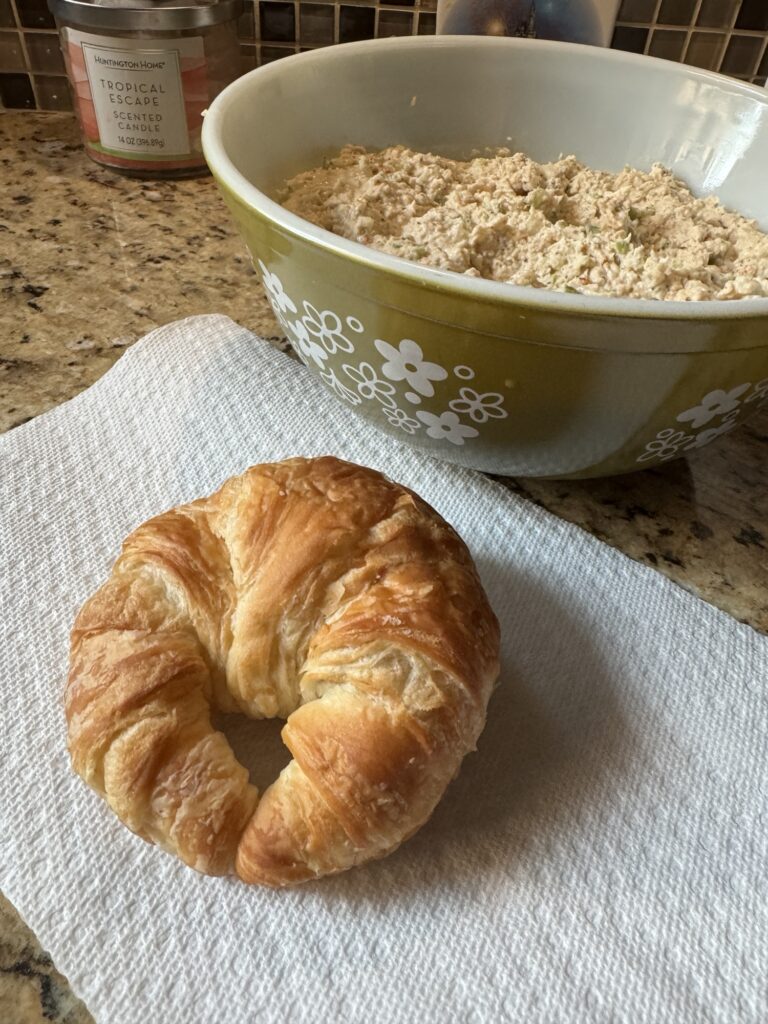 A croissant on a paper towel next to a bowl of chicken salad