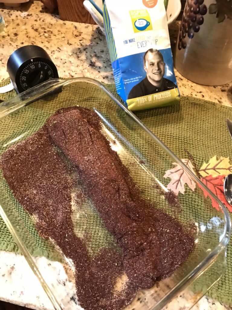 A pork tenderloin in a glass baking dish covered in spice rub. There is a kitchen timer and a package of Aspire Coffee. 
