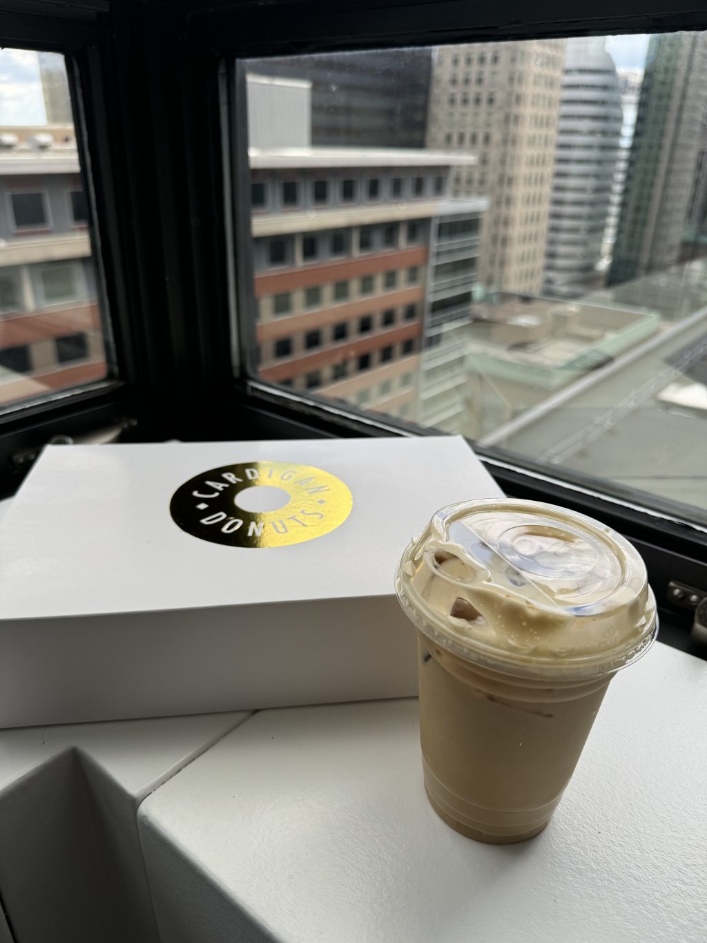 An iced coffee and a closed box of Cardigan Donuts on the window sill of my hotel room. 