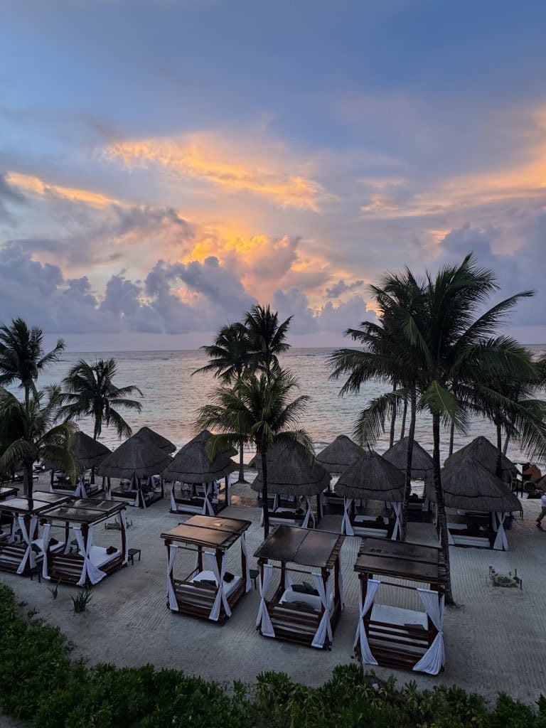 A view of the beach, with palm trees and beach beds under straw huts, at sunrise.