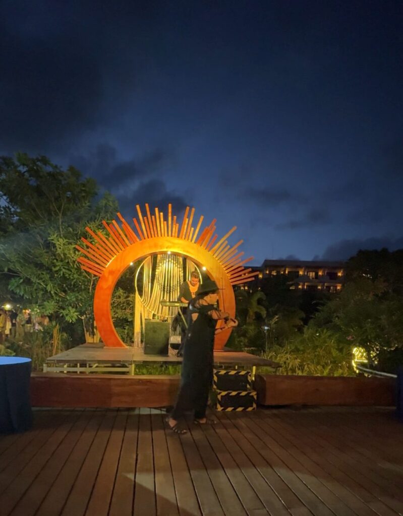 A performer playing violin standing on a wooden stage. Behind them is a keyboard player on a raised section of the stage under an orange circular sculpture at night.