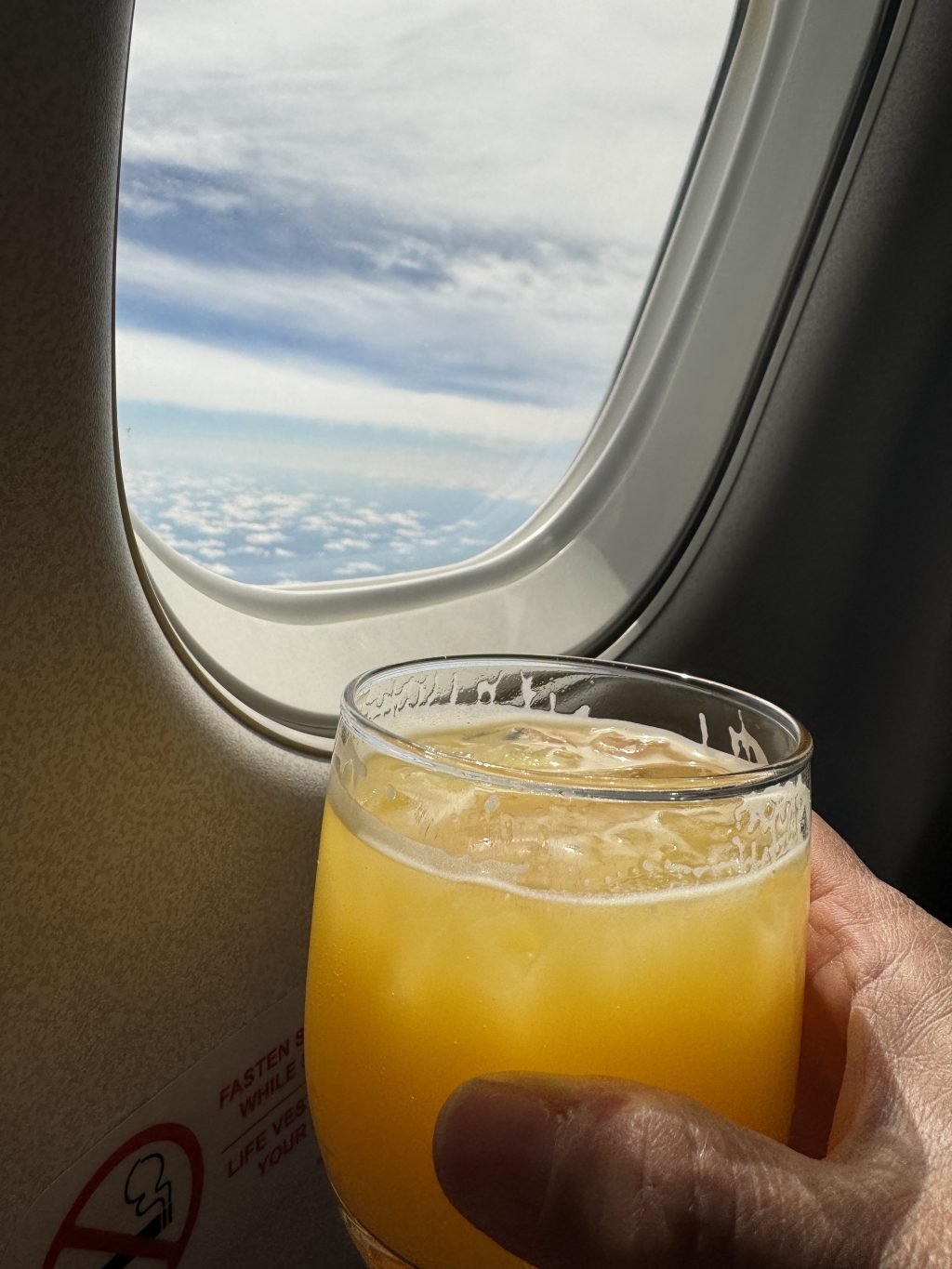 A glass of orange juice being held by a plane window in the air.