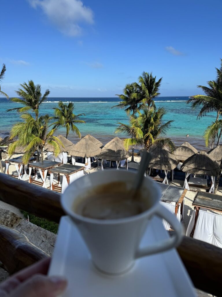 A cup of espresso with a spoon in it. being held in front of a beautiful beach view.