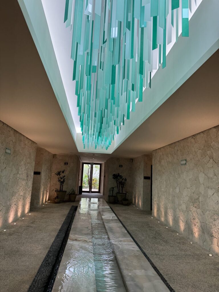 A hall in the Secrets Akumal resort. The walls are stone and there are glass decorations hanging from the ceiling and a canal of shallow water in  the middle of the hall. 