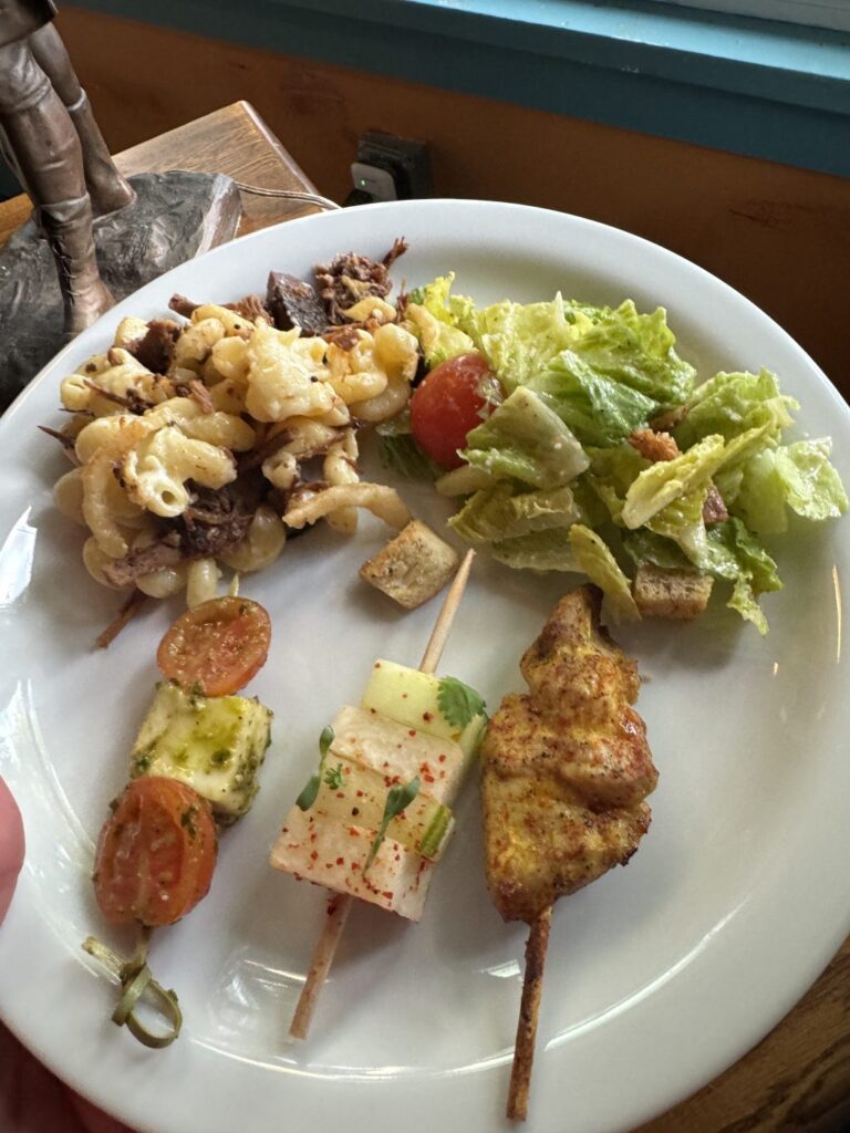 Skewers of chicken and vegetables alongside salad and meaty mac and cheese on a white plate.