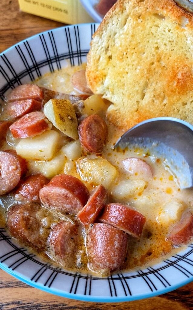 A close up of a spoon going into a bowl of frankfurter and potato soup with a crust of bread.