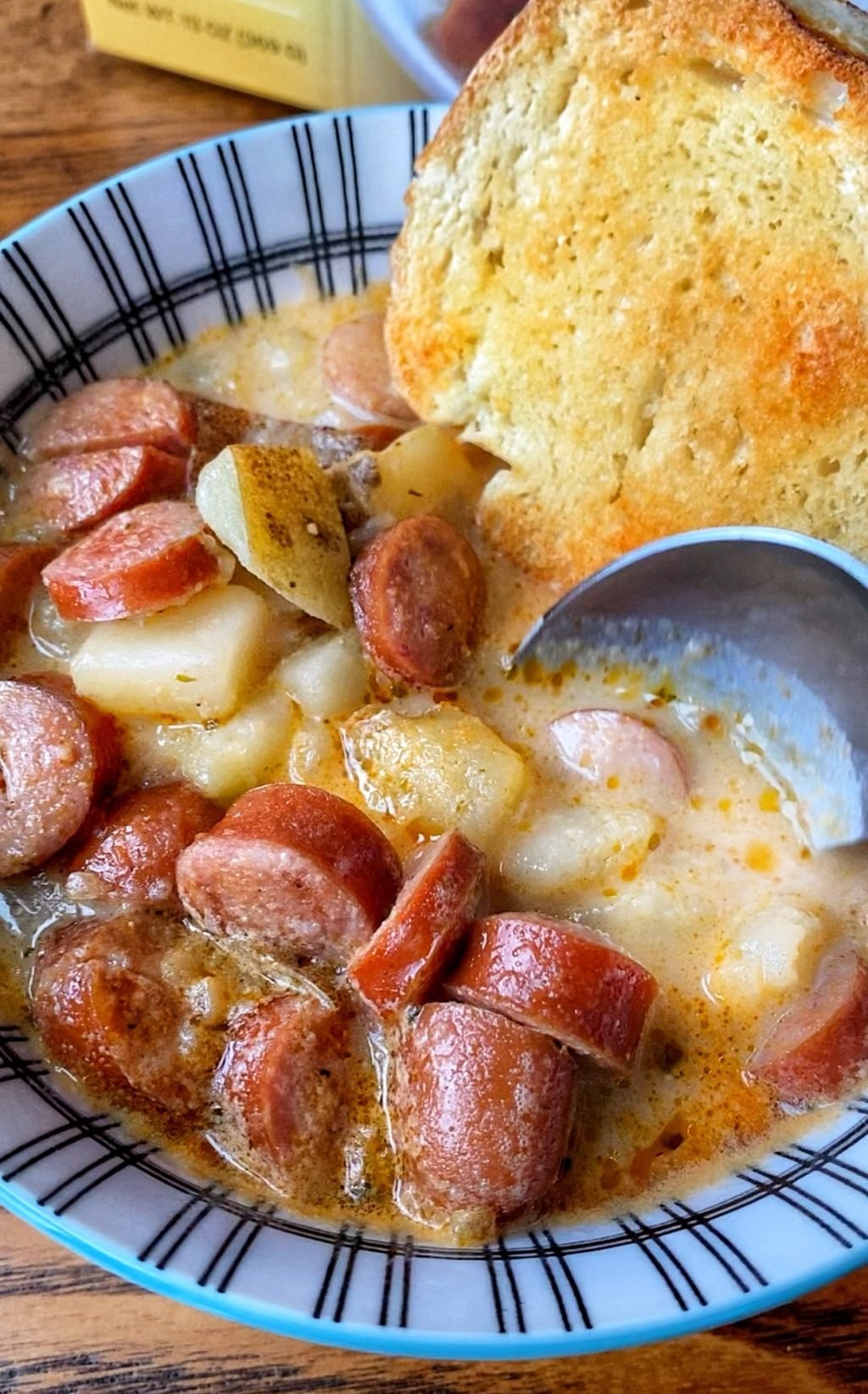 A close up of a spoon going into a bowl of frankfurter and potato soup with a crust of bread.
