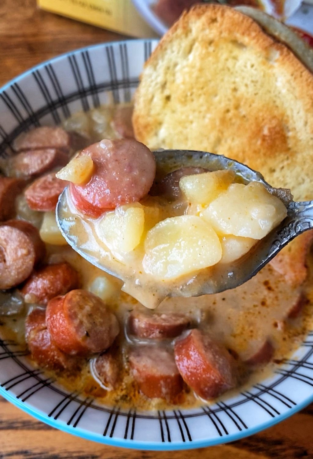A close up of a spoon of frankfurter and potato soup over a bowl of more soup with a crust of toasted bread.