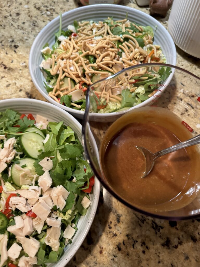 Two bowls of undressed Chinese Chicken Salad and a measuring cup of brown peanut dressing with a spoon in it.