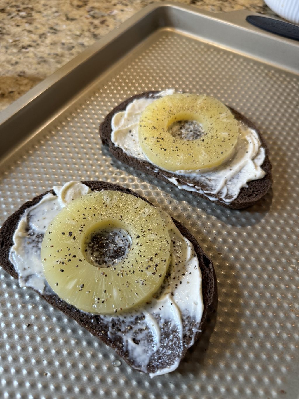 Two pieces of pumpernickel bread with mayonnaise, a pineapple ring, and pepper on top.