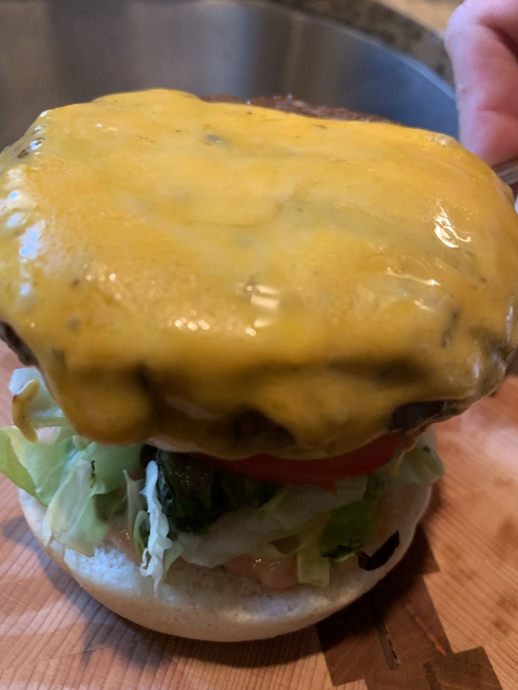 Cheese melted over a burger held on a utensil above a bun with lettuce and tomato on it.