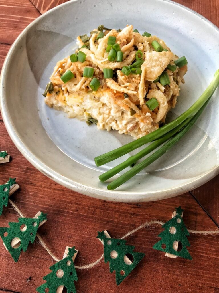 A slice of crockpot potatoes covered in green onions and crispy onions on a blue plate next to small felt Christmas trees.