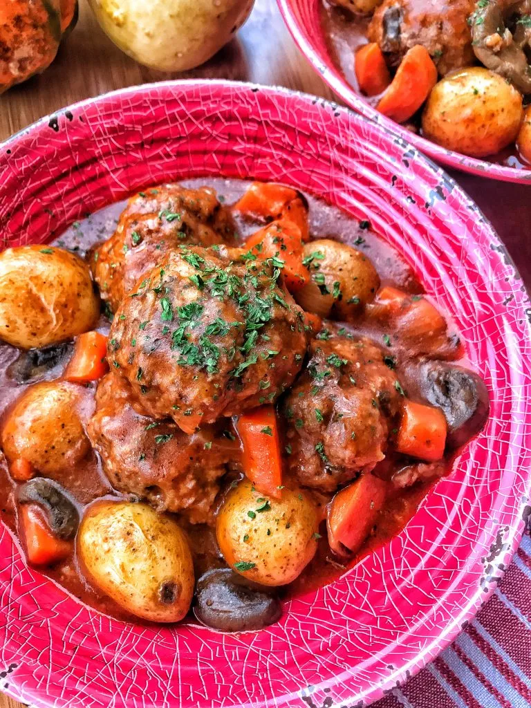 A red bowl with pot roast meatballs and gravy over carrots, mushrooms, and potatoes.