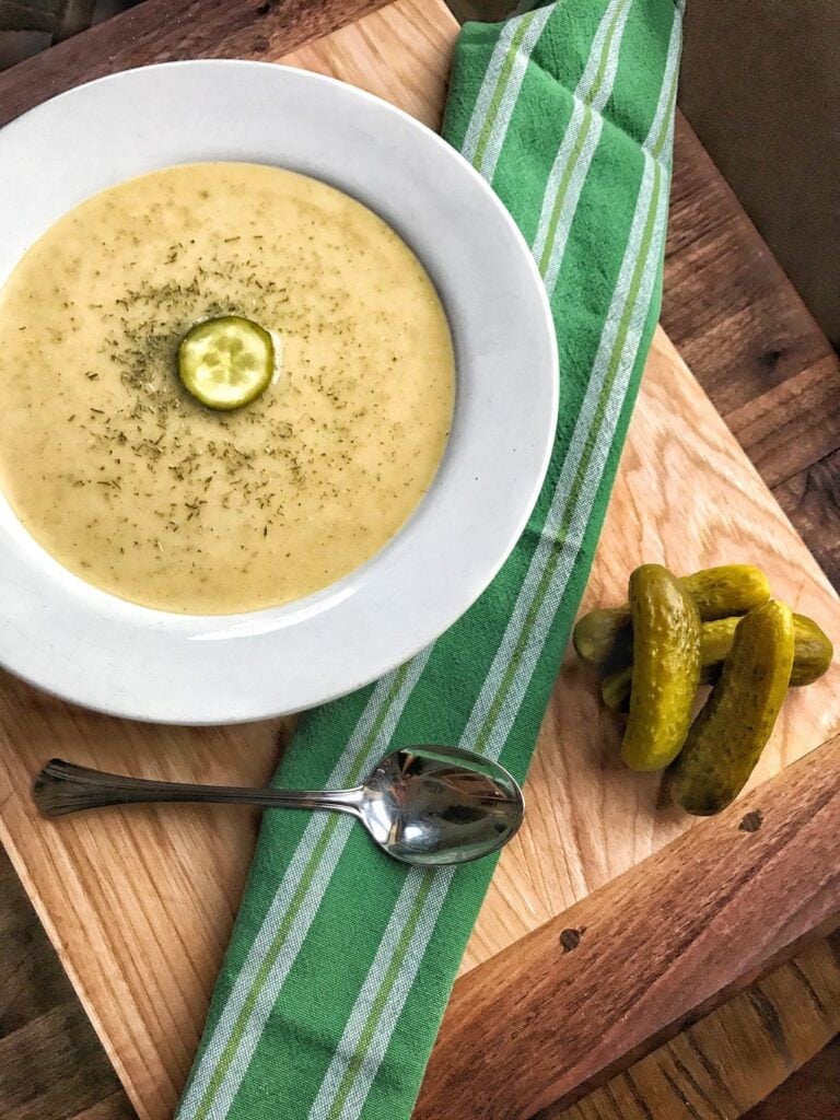 A bowl of dill pickle soup on a wooden cutting board next to some pickles and a spoon.