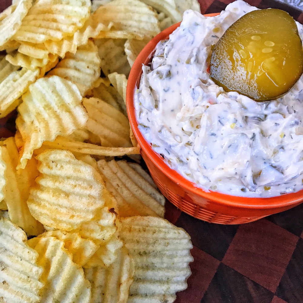 White dip with a pickle chip in it surrounded by potato chips.