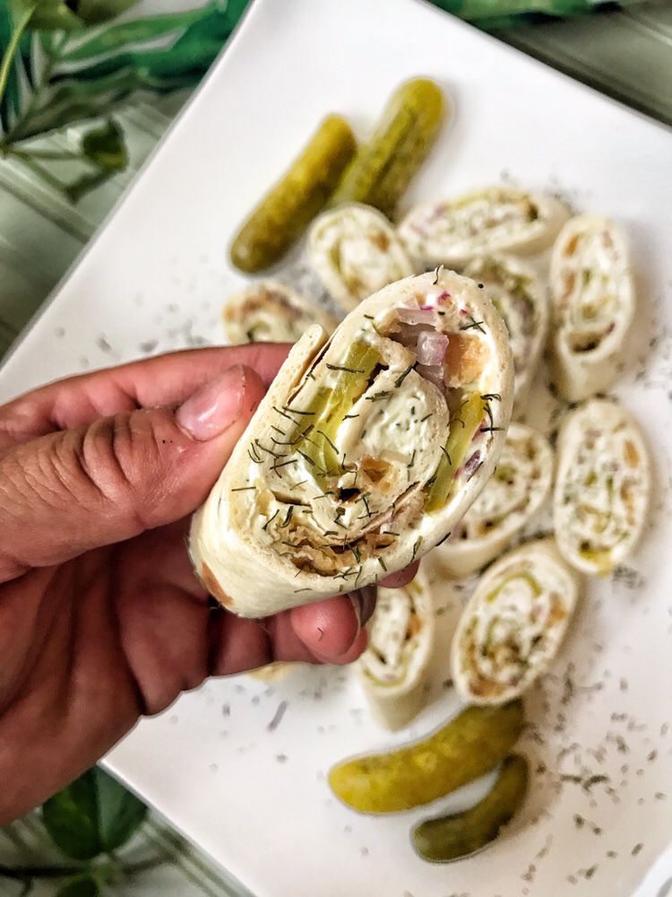 A close up of a pickle pinwheel held above a plate of more pinwheels.