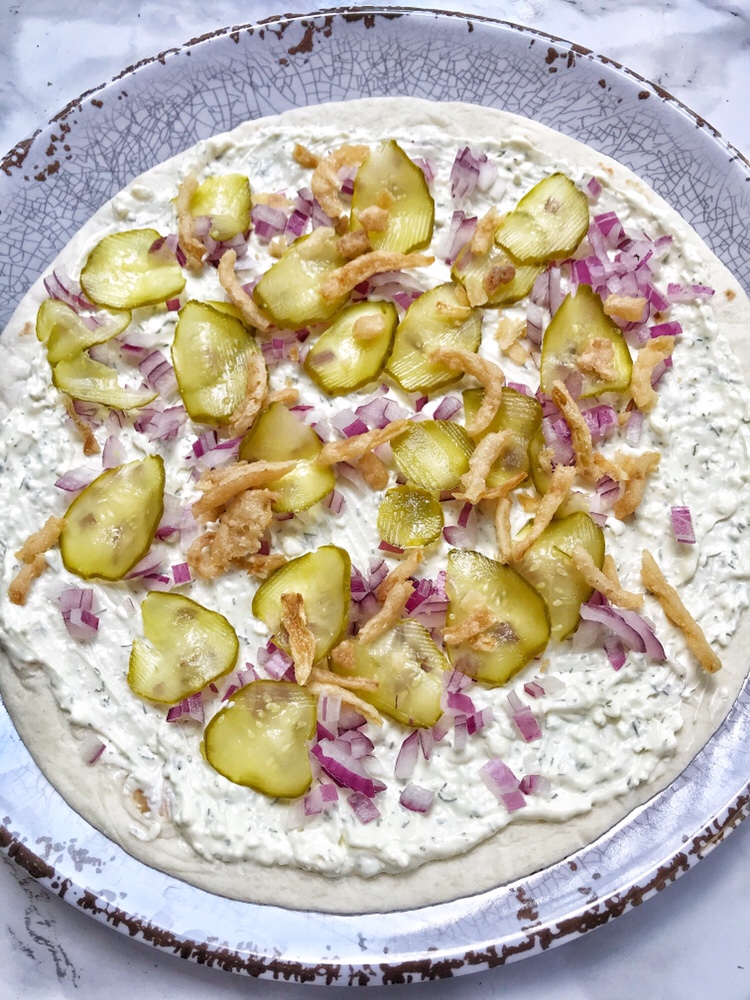 Pickle slices, crunchy onions, diced red onions, and cream cheese spread over an open tortilla on a plate.