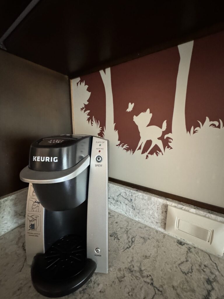 A Keurig coffee machine on a hotel counter. Behind it a white outline of Bambi in the forest is on the wall.