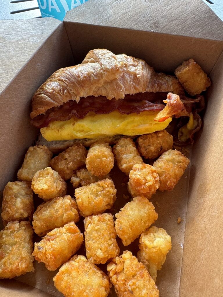 A cardboard box with a bacon, egg, and cheese croissant and tater tots.