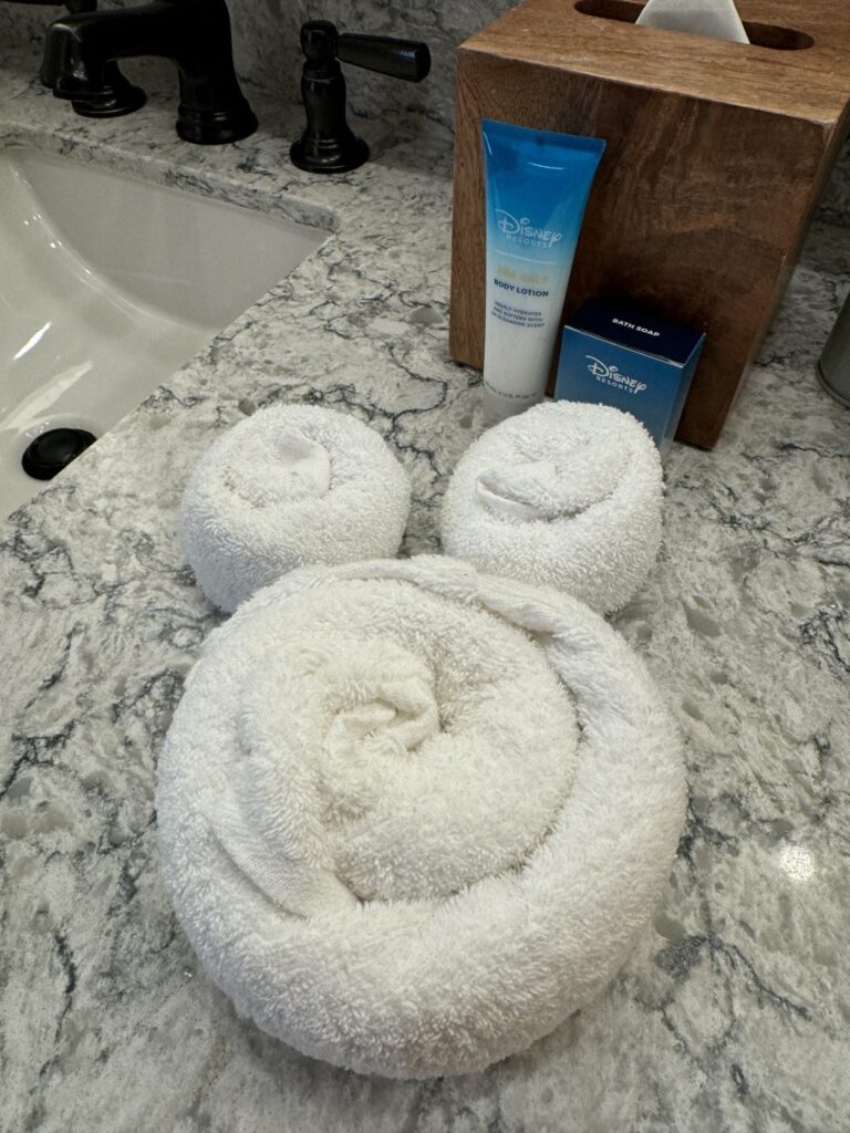 Hand towels rolled into the shape of the iconic Mickey Mouse logo on a bathroom counter next to a tissue box and Disney toiletries. 