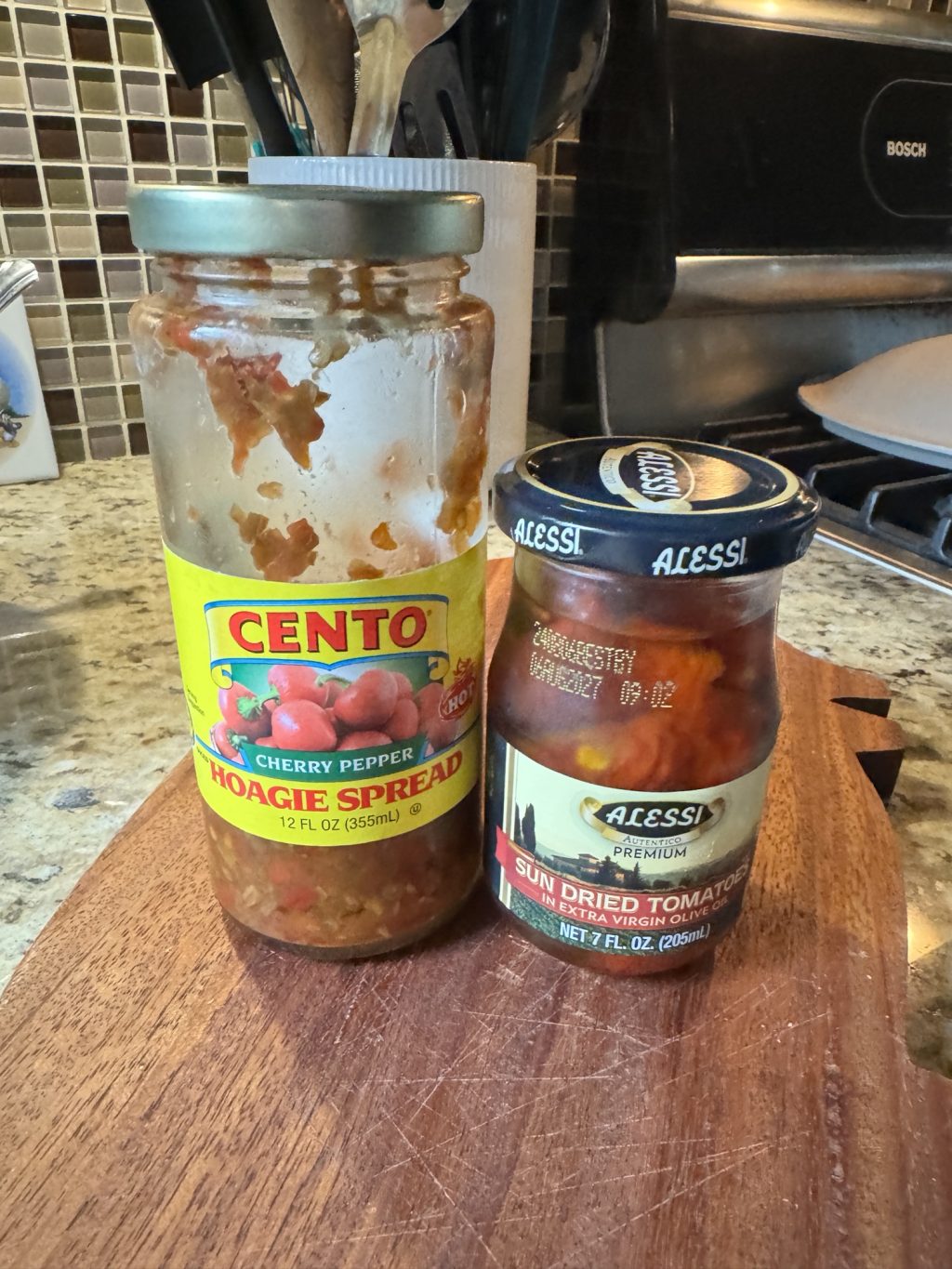 A jar of cherry pepper hoagie spread next to a jar of sun dried tomatoes in extra virgin olive oil.
