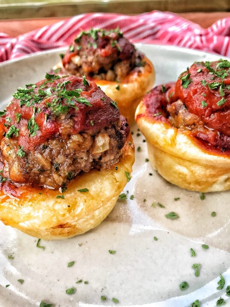 Three meatball cups on a plate. They are meatballs in dough cups with a little tomato sauce on top.