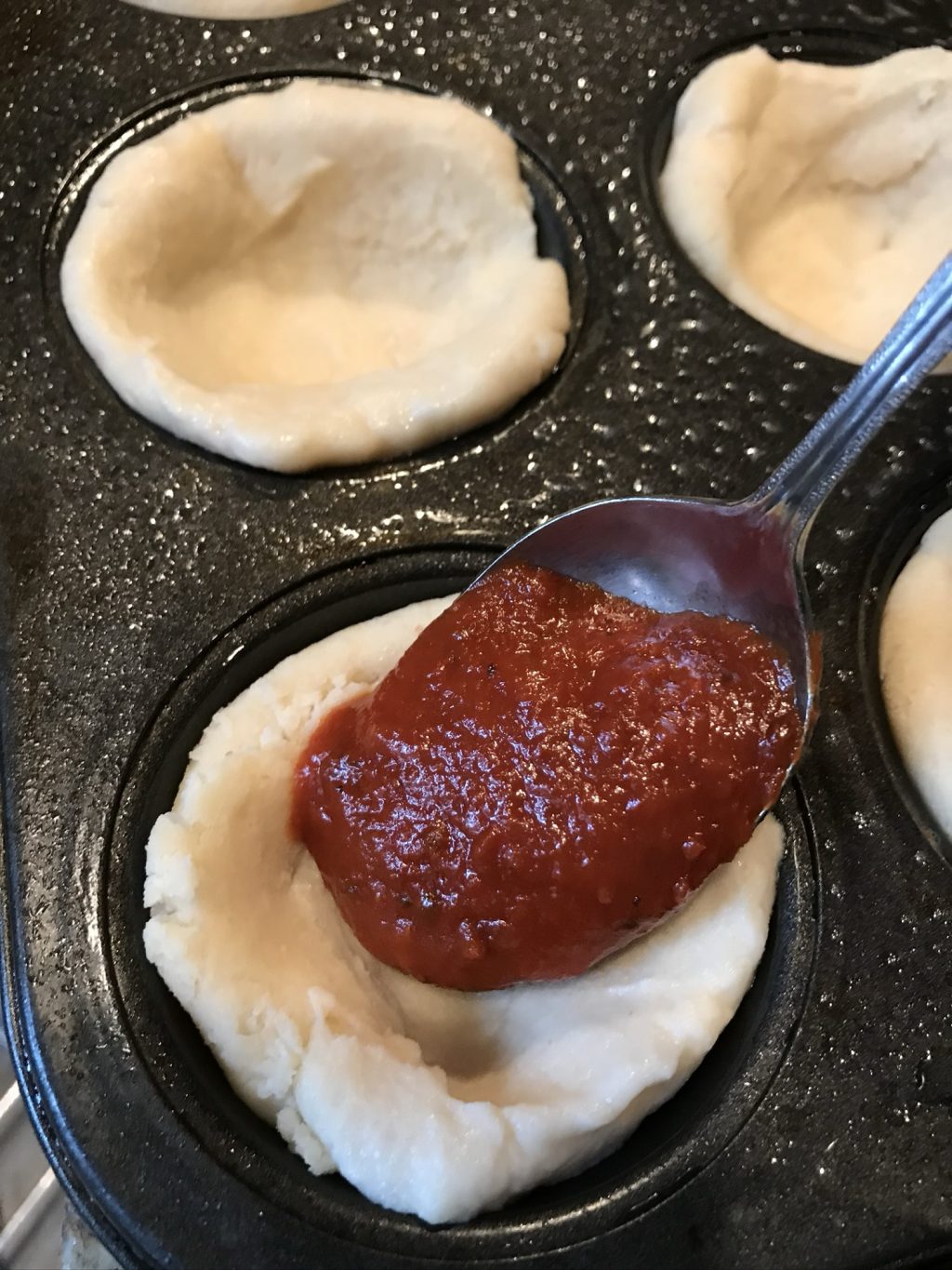 Pizza sauce being spooned into one of the dough filled muffin tin cups.