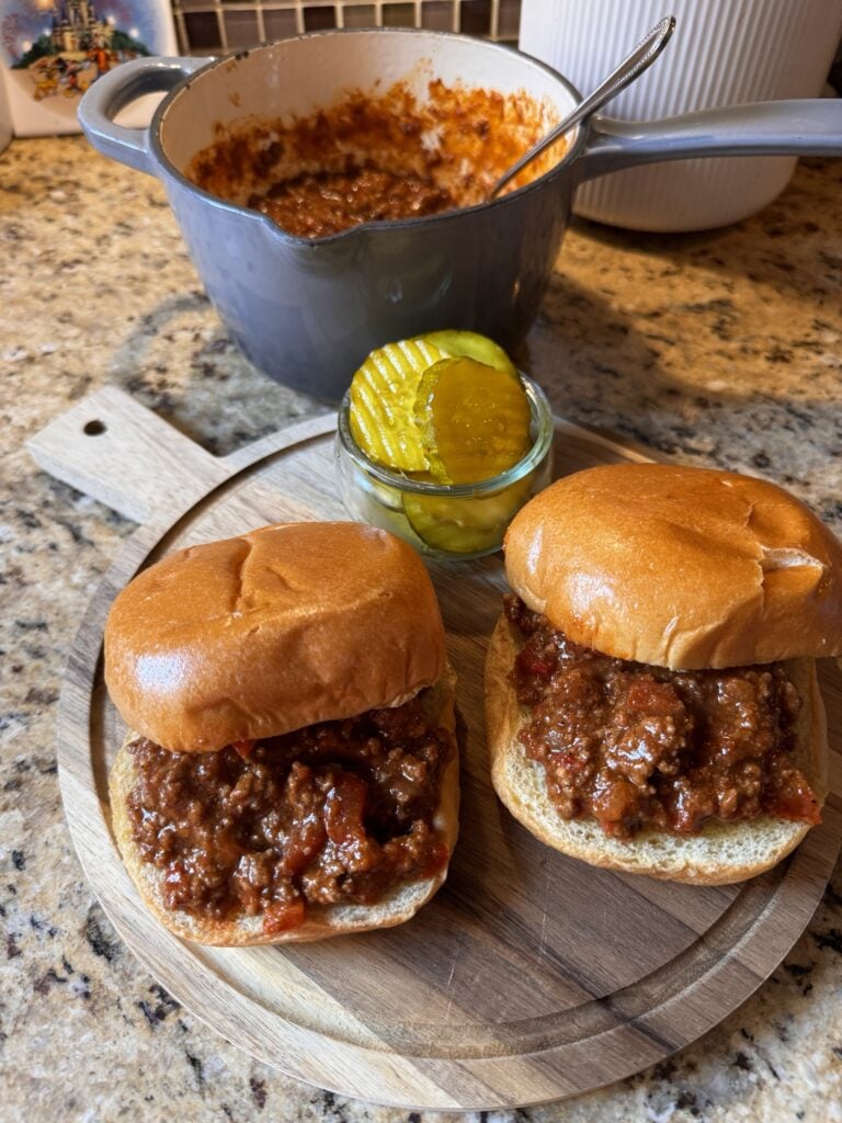 Two BBQ beef sandwiches on a cutting board with a small cup of pickle chips from above.
