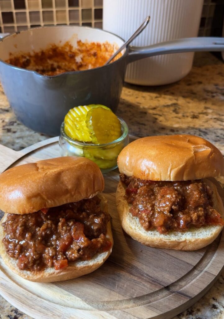 Two BBQ beef sandwiches on a cutting board with a small cup of pickle chips.