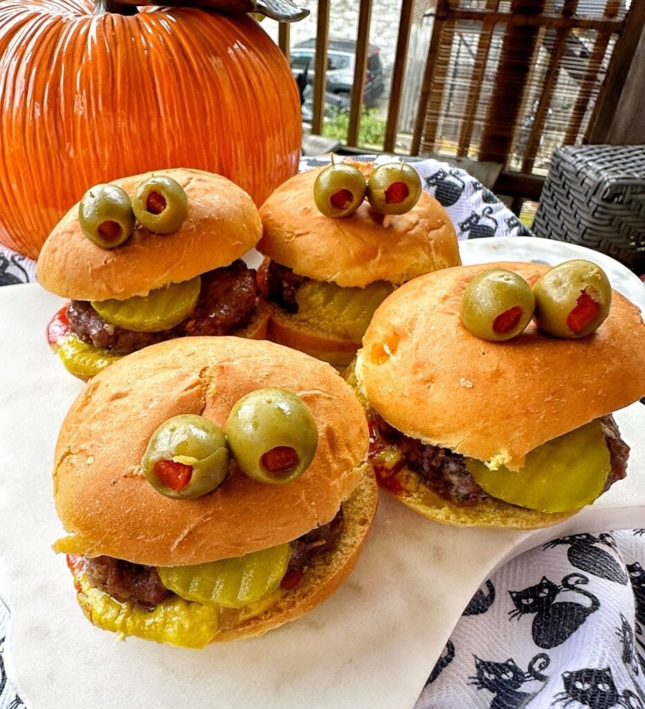 Four burgers with olive "eyes" in front of a pumpkin.