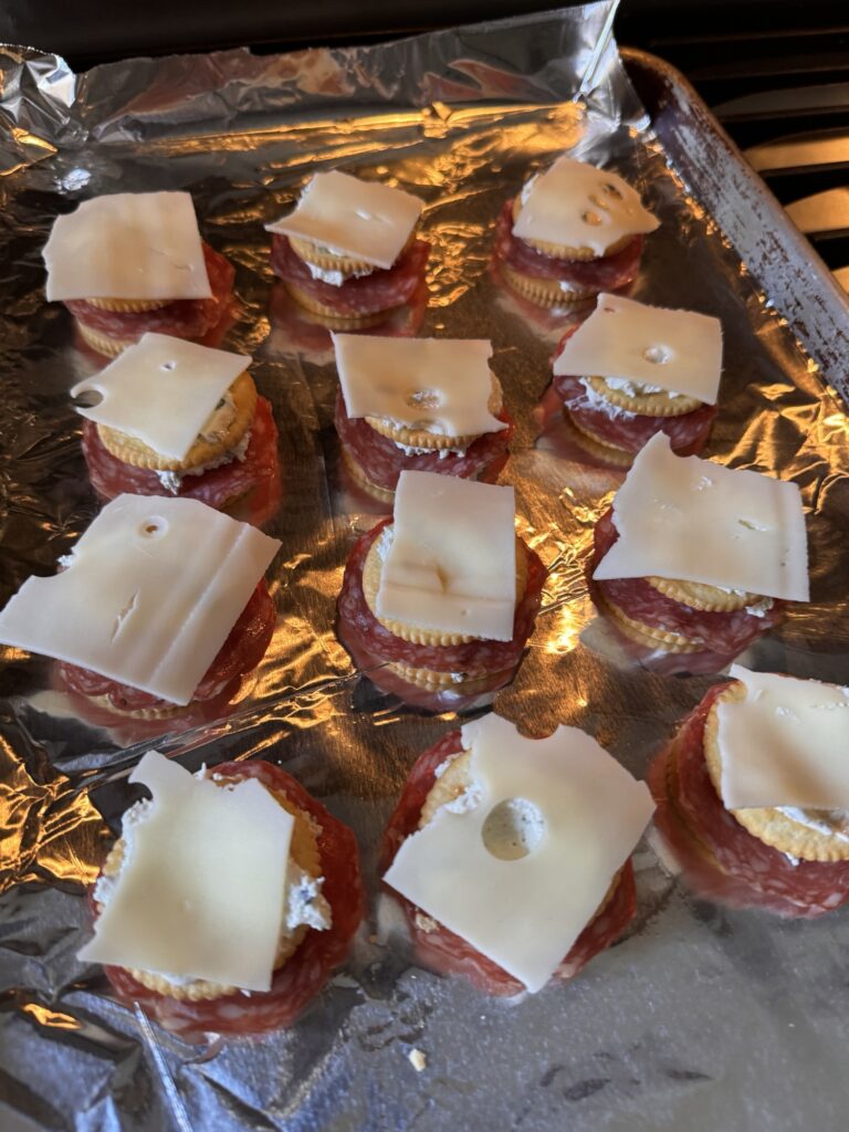 Crackers spread out on a foil covered baking sheet topped with salami, another cracker, and cheese.