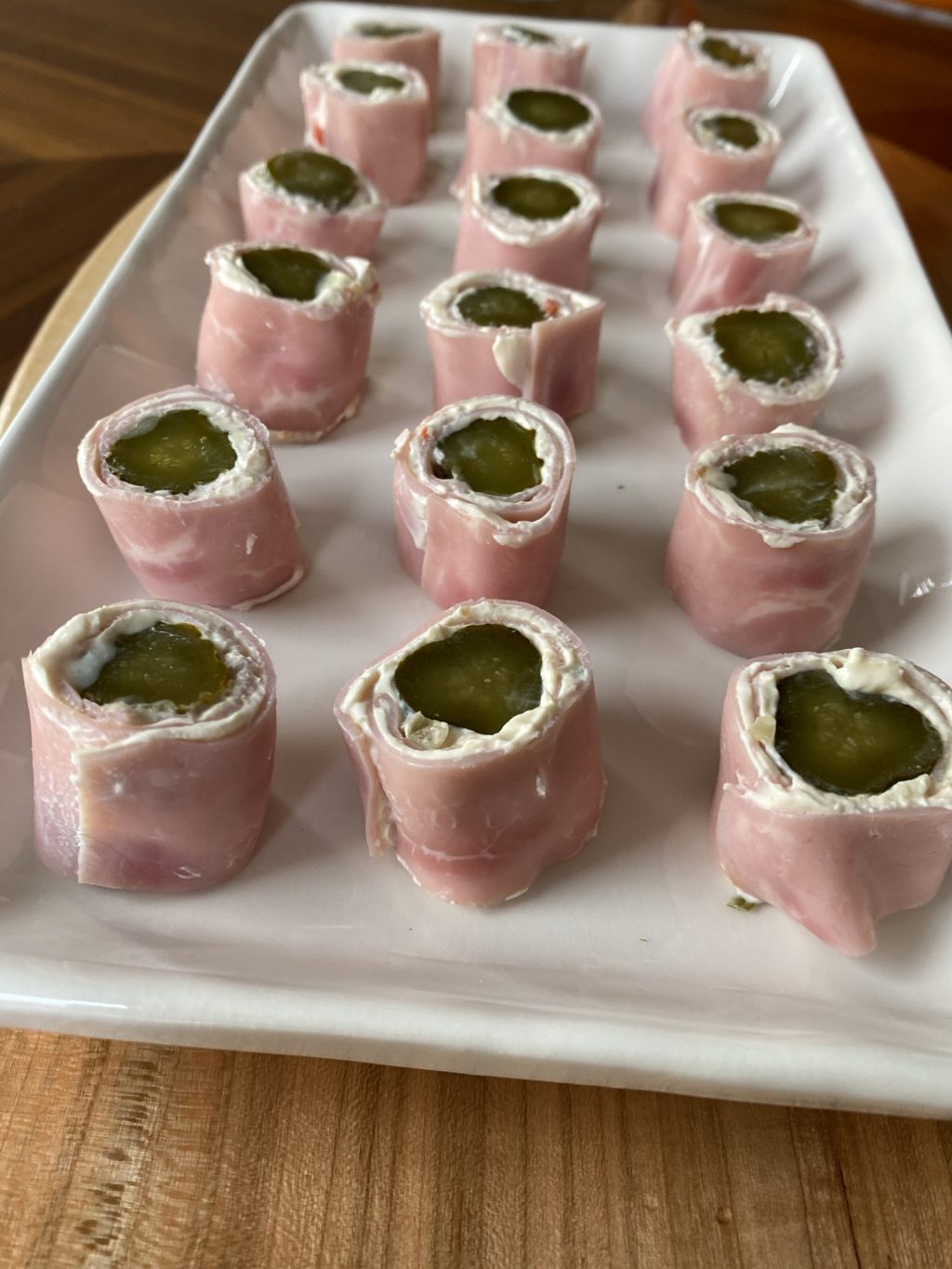 A white rectangular plate with bites of ham wrapped around cream cheese covered pickle chunks.