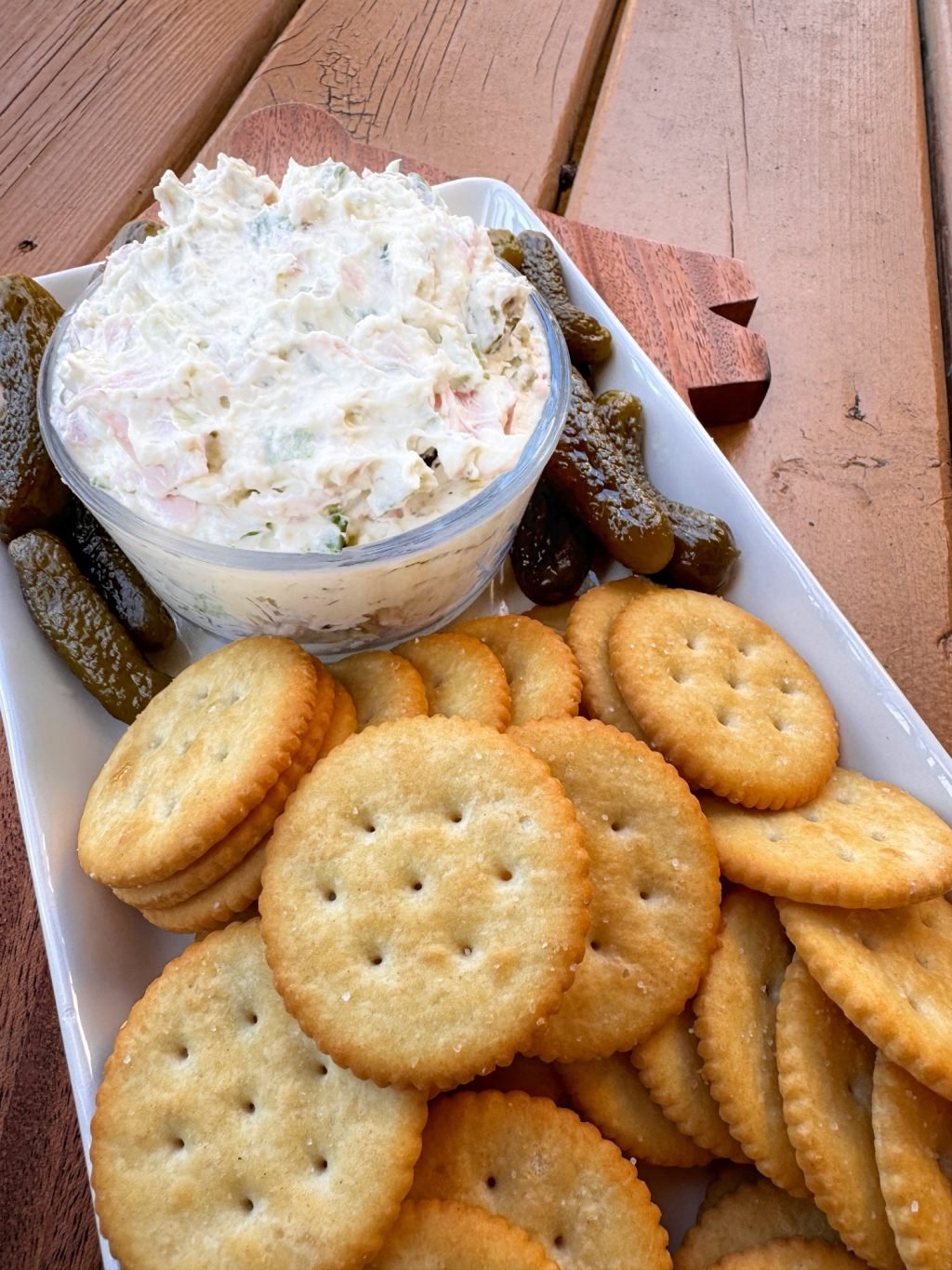 Another angle of a white dip with chunks of ham and pickles on a rectangular plate with small pickles and butter crackers.