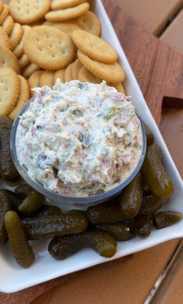 A close up of white dip with chunks of ham and pickles on a rectangular plate with small pickles and butter crackers.