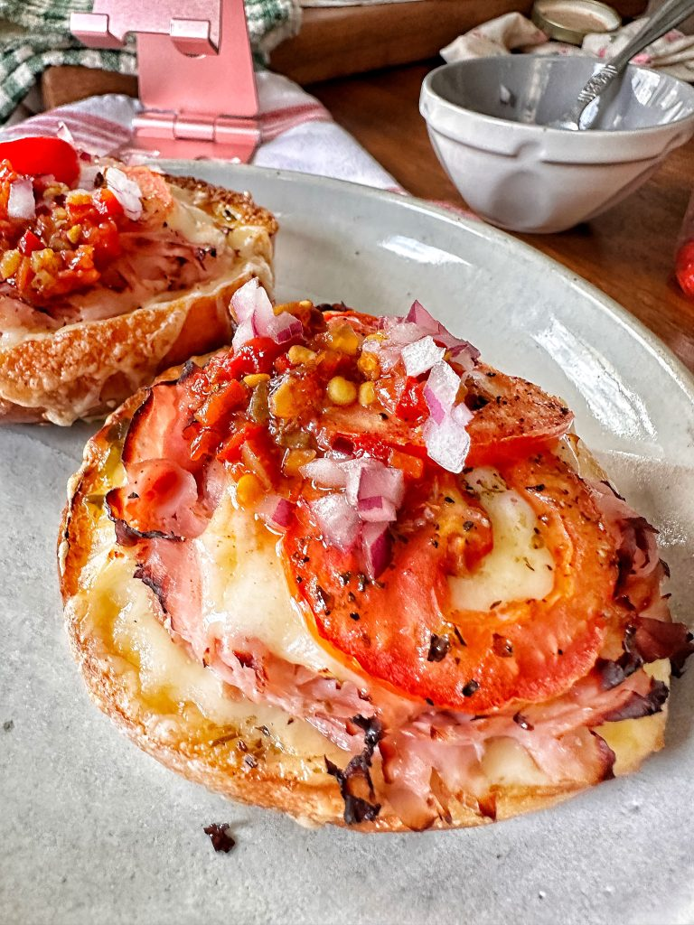 Two open faced toasty ham sandwiches topped with tomato sliced and diced onion on a plate.