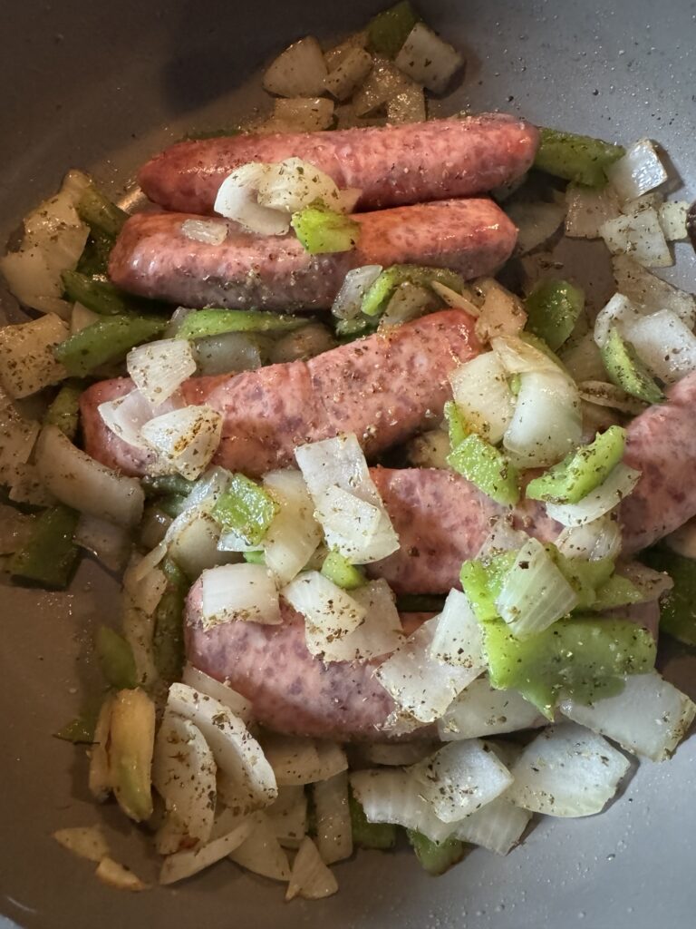 Sausages cooking in a pan with chopped onions, pepper pieces, and seasoning.