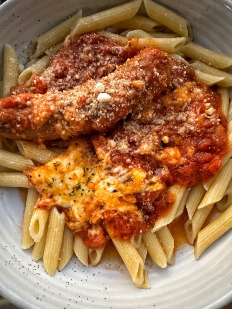 Penne pasta in a white bowl topped with tomato sauce, cheese, and sausage.