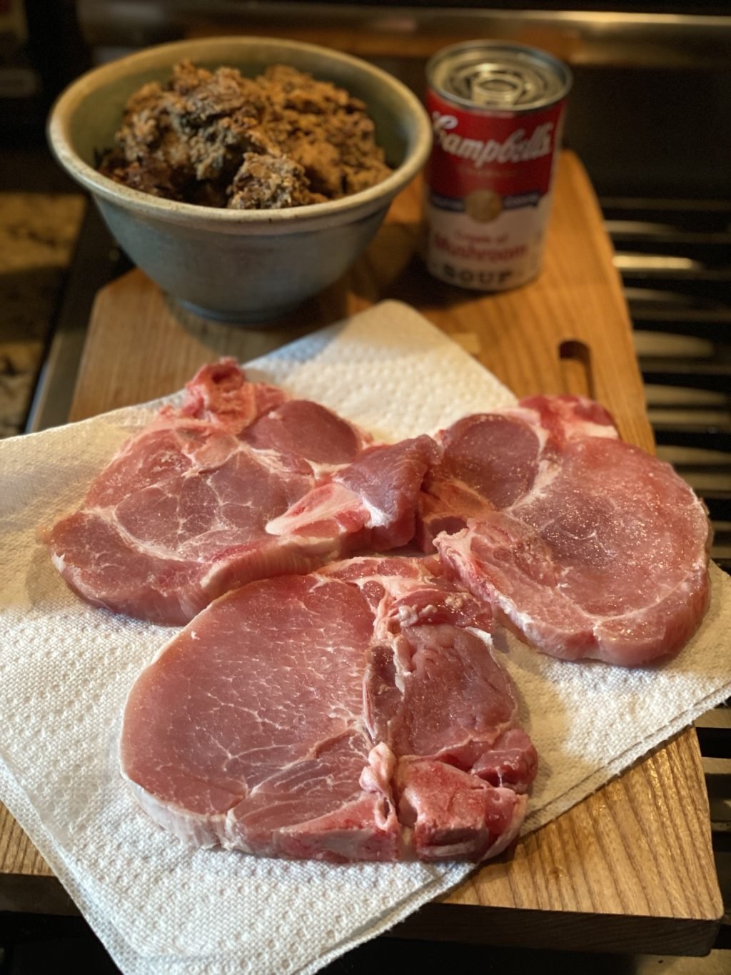 Three raw pork chops on a paper towel on a wooden cutting board in front of a bowl of stuffing and a can of Campbell's Mushroom Soup.